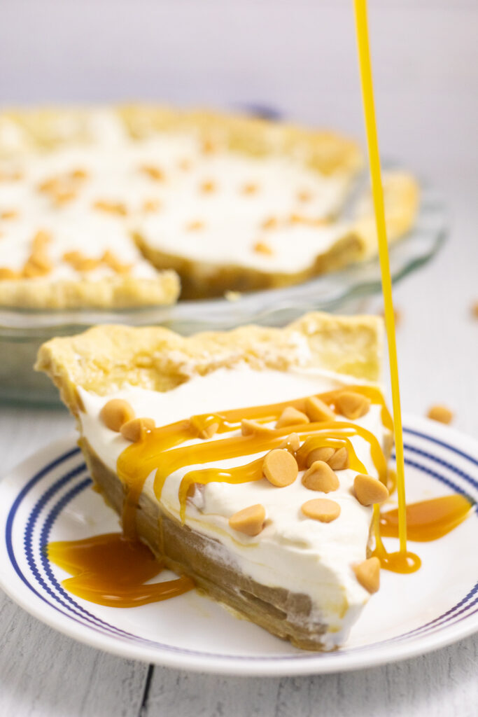 Butterscotch sauce being drizzled onto homemade butterscotch pie on a small white plate with a rustic white wood background.