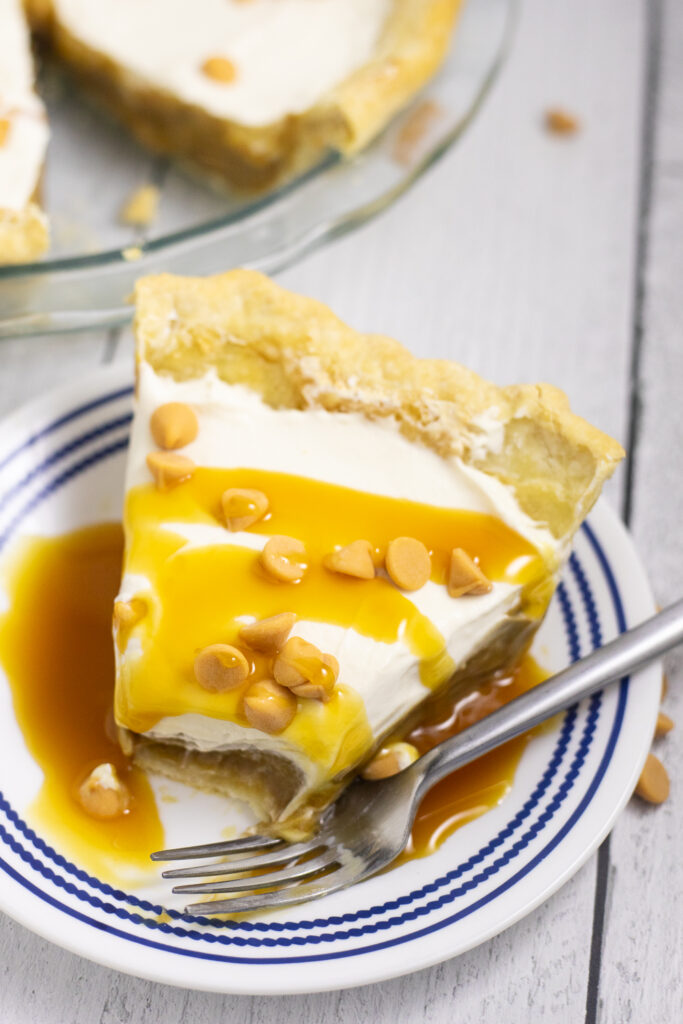 Overhead view of a slice of butterscotch pie on a small plate with a fork and a bite missing. The pie is drizzled with butterscotch sauce and topped with butterscotch chips.