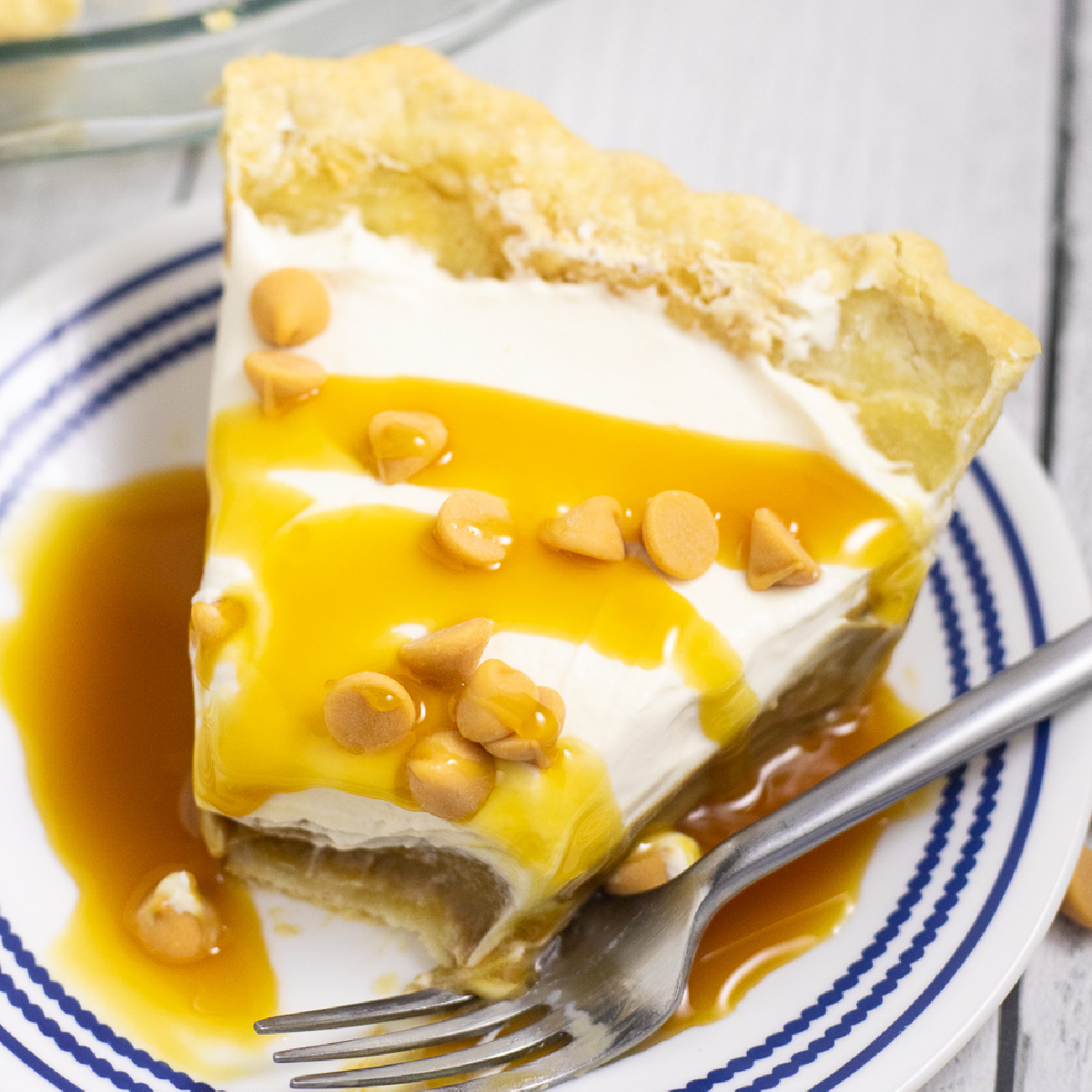 Overhead view of a slice of butterscotch pie on a small plate with a fork and a bite missing. The pie is drizzled with butterscotch sauce and topped with butterscotch chips.