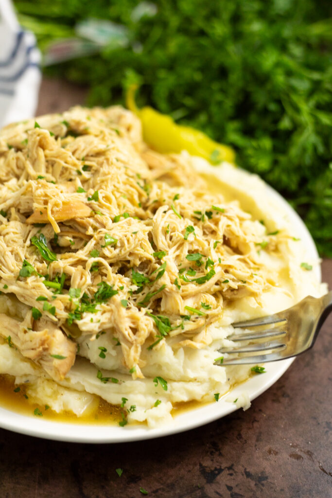 Crockpot Mississippi Chicken on top of mashed potatoes topped with fresh parsley with a fork taking a bite out of it wit fresh parsley behind.