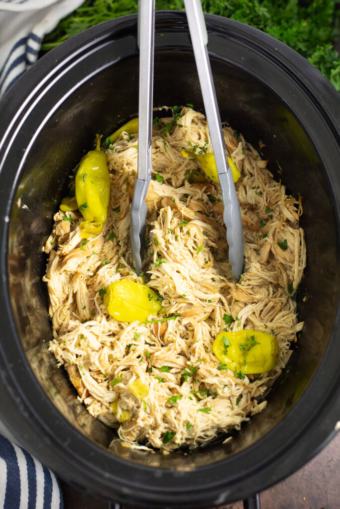 Overhead view of Crockpot Mississippi Chicken in a slow cooker with pepperoncinis and parsley on top and tongs in the crock.
