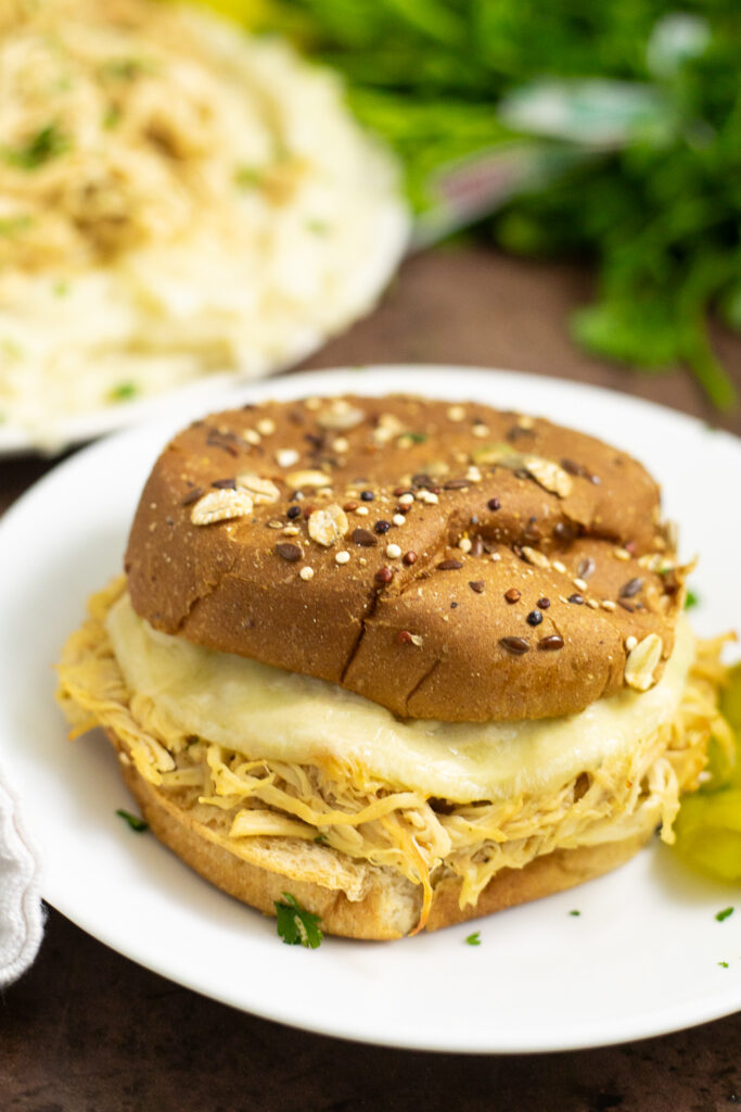 Crockpot Mississippi chicken on a bun with melted cheese. The sandwich is on a small white plate with mashed potatoes and a bunch of fresh parsley behind.
