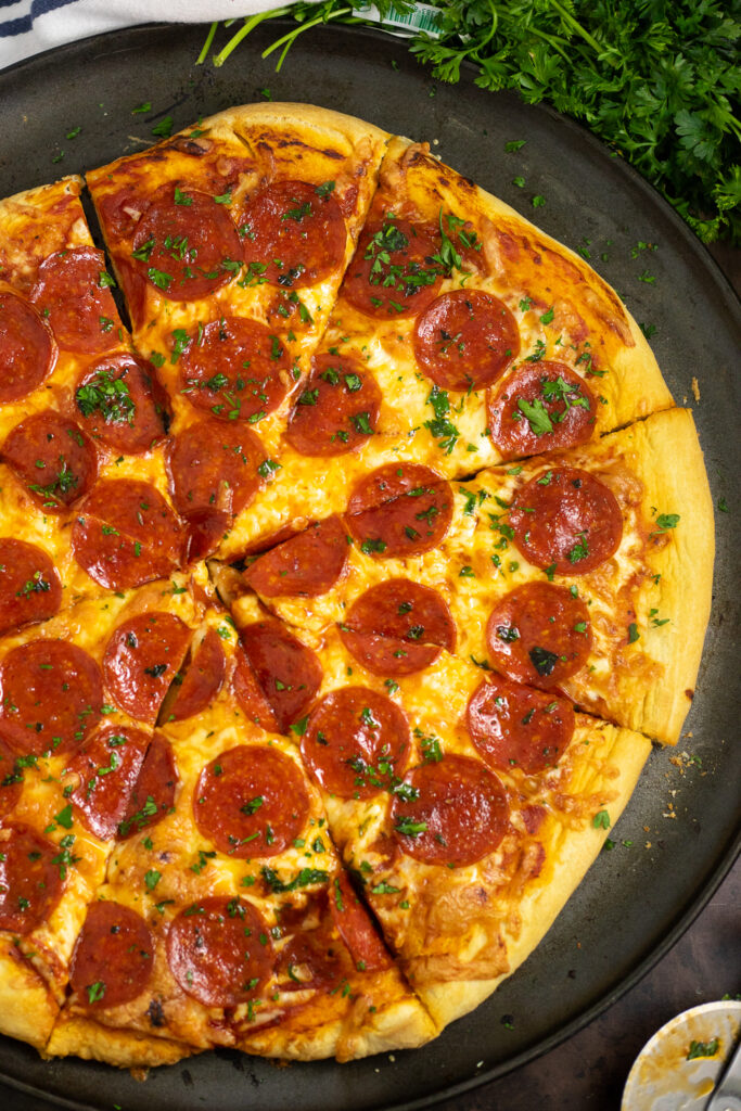 Overhead view of a baked homemade pepperoni pizza with homemade pizza crust on a round pizza pan next to a bunch of fresh parsley.