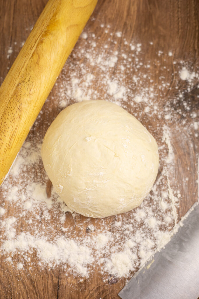 Overhead view of a ball of homemade pizza dough on a floured wooden surface next to a wooden rolling pin and a bench scraper.