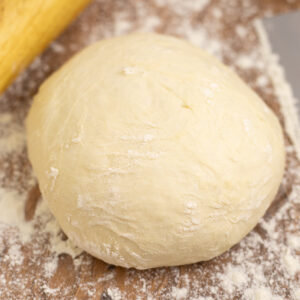 A dough of homemade pizza dough on a floured wooden surface next to a wooden rolling pin.