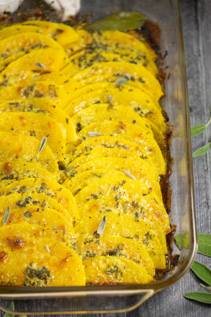 Baked butternut squash topped with garlic sage butter and bubbly parmesan in a glass casserole dish on a rustic wood background surrounded by fresh sage leaves.