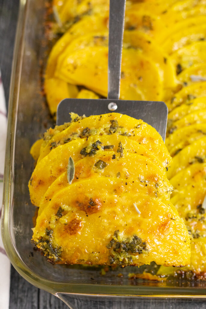 Spatula holding up three slices of baked butternut squash covered in garlic sage butter and topped with bubbly parmesan cheese.