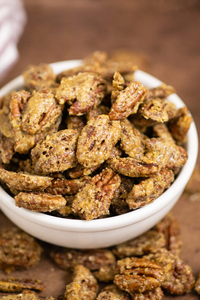 Candied pecans in a medium ceramic white bowl surrounded by more candied pecans on a distressed warm brown background.