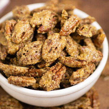 Candied pecans in a medium ceramic white bowl surrounded by more candied pecans on a distressed warm brown background.