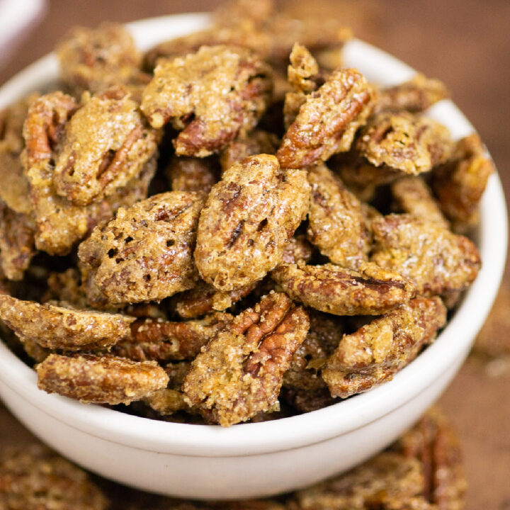 Candied pecans in a medium ceramic white bowl surrounded by more candied pecans on a distressed warm brown background.