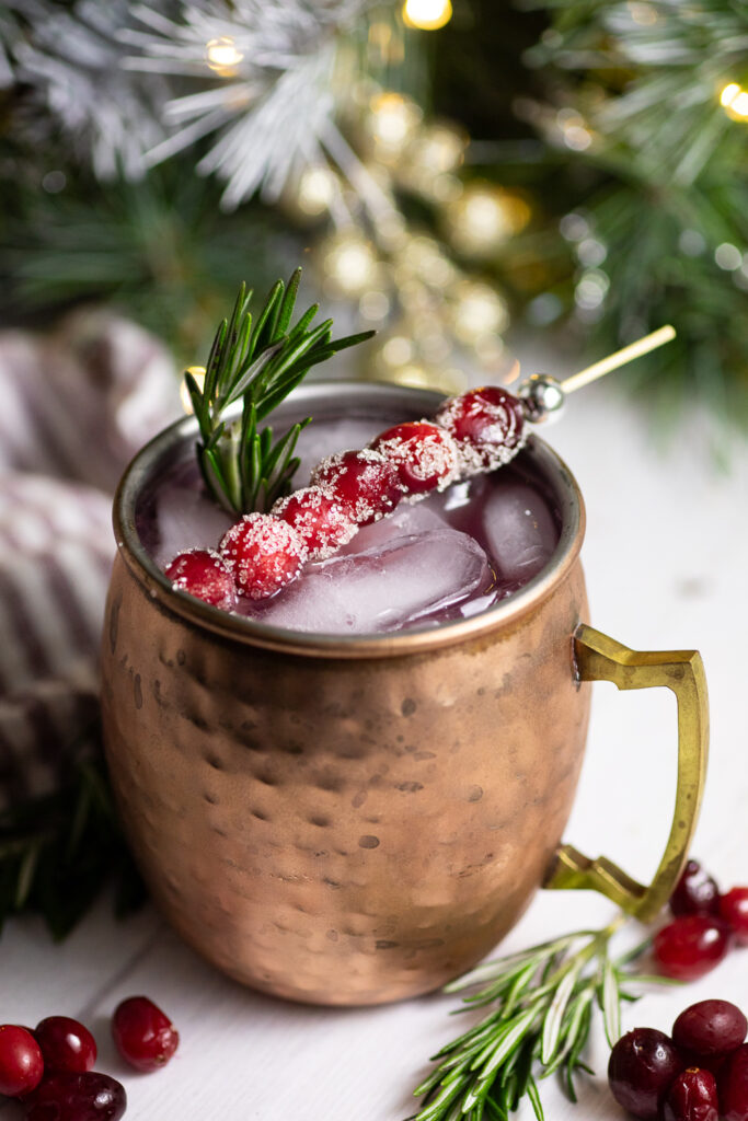Cranberry Moscow Mule in a copper mug with ice garnished with sugared cranberries on a toothpick and a sprig of rosemary, with more rosemary and cranberries around it on a white background and a red striped linen napkin and a Christmas garland behind.