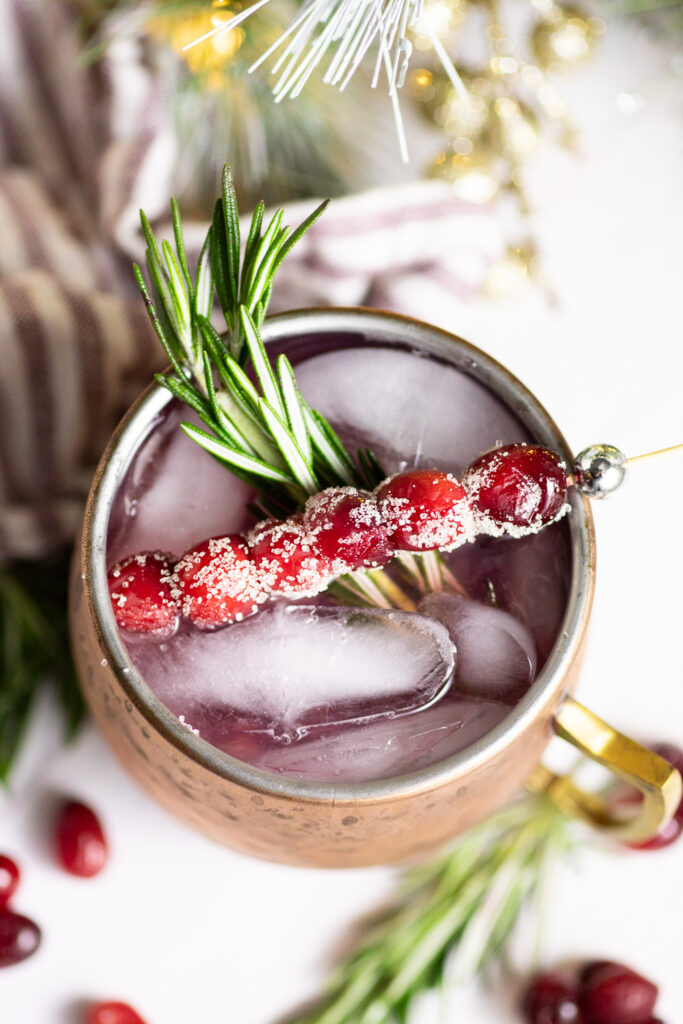 Overhead view of a Cranberry Moscow Mule in a copper mug with ice garnished with sugared cranberries on a toothpick and a sprig of rosemary, with more rosemary and cranberries around it on a white background.
