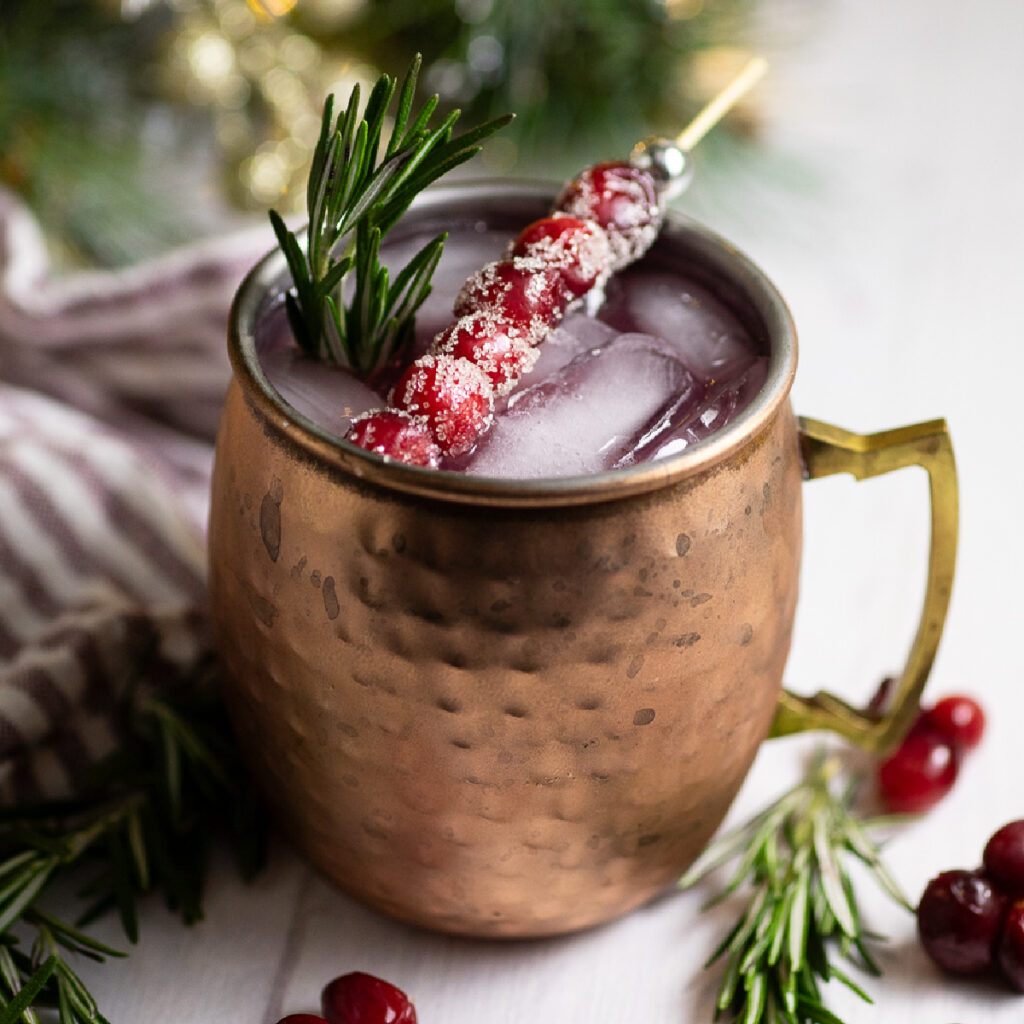 Cranberry Moscow Mule in a copper mug with ice garnished with sugared cranberries on a toothpick and a sprig of rosemary, with more rosemary and cranberries around it on a white background and a red striped linen napkin and a Christmas garland behind.