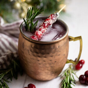 Cranberry Moscow Mule in a copper mug with ice garnished with sugared cranberries on a toothpick and a sprig of rosemary, with more rosemary and cranberries around it on a white background and a red striped linen napkin and a Christmas garland behind.