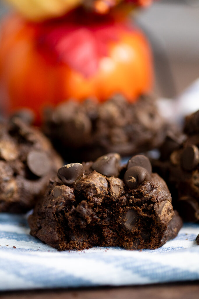 Double chocolate pumpkin cookie with a bite taken out sitting on a white and blue striped linen with more cookies behind.