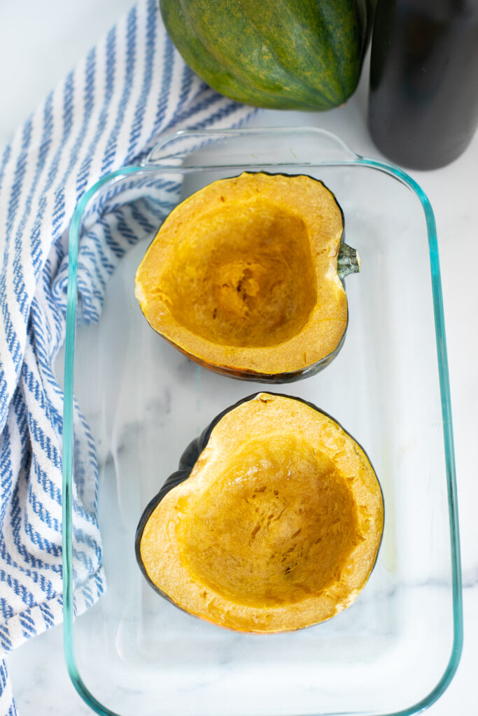Overhead view of an acorn squash cut in half with the seeds scooped out in a small glass baking dish on a white background with a whole acorn squash and a bottle of olive oil behind.
