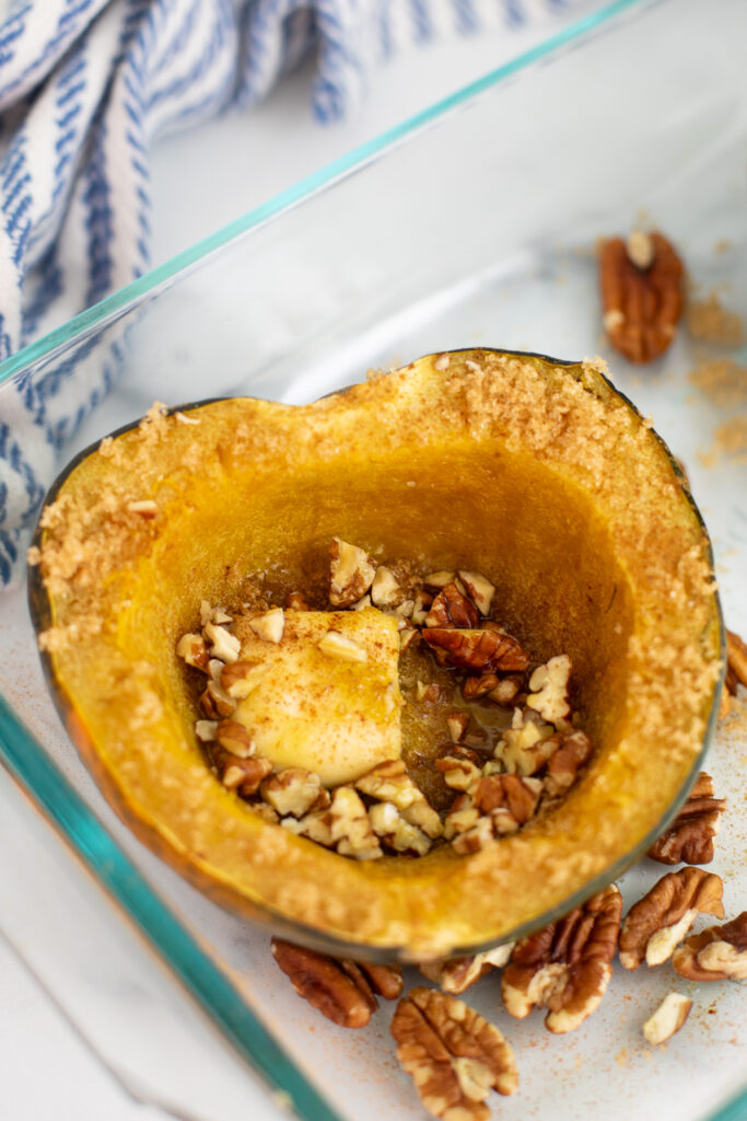 Half of a cooked microwave acorn squash topped with a pat of butter, brown sugar, and chopped pecans in a glass baking dish.