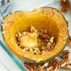 Half of an acorn squash in a glass dish topped with a pat of butter, brown sugar, and chopped pecans.