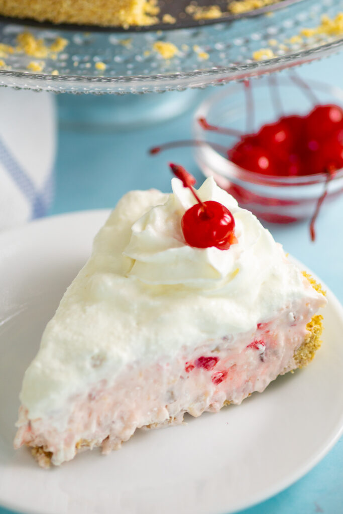 A piece of millionaire pie topped with whipped cream and a maraschino cherry on a small white plate with a bright blue background and a small glass bowl of cherries behind.