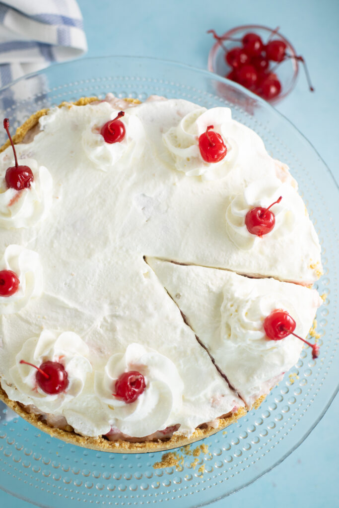 Overhead view of millionaire pie topped with whipped cream and maraschino cherries with a slice cut out on a bright blue background with a bowl of maraschino cherries and a linen napkin next to it.