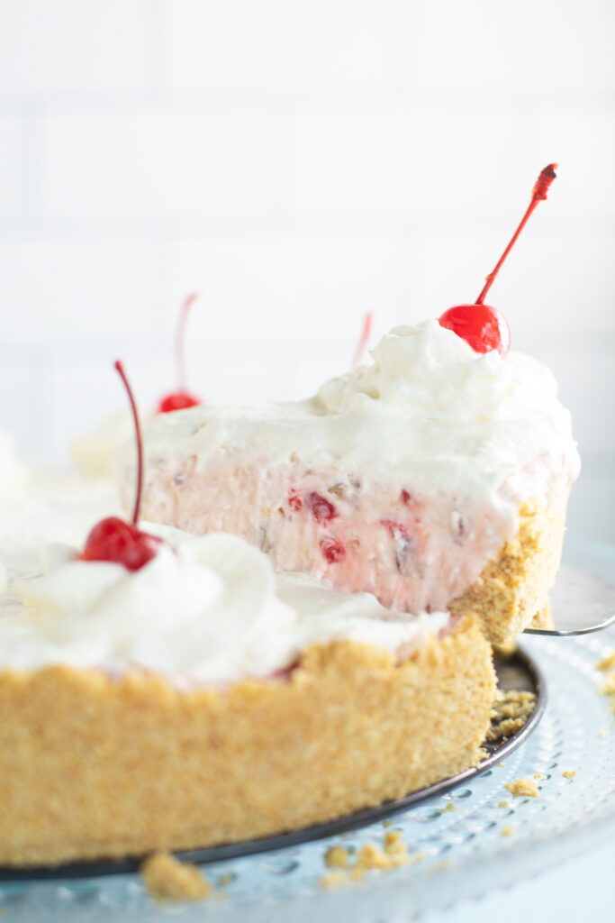 A slice of pie topped with a maraschino cherry and whipped cream being lifted from the full pie.