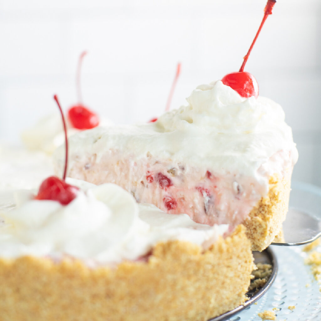 A slice of pie topped with a maraschino cherry and whipped cream being lifted from the full pie.