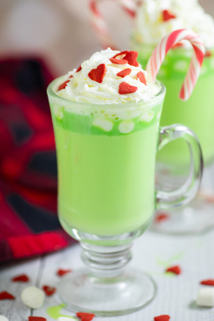 Green Grinch hot chocolate in a glass mug with a stem. The hot chocolate is topped with whipped cream, small red heart sprinkles, and a peppermint candy cane.