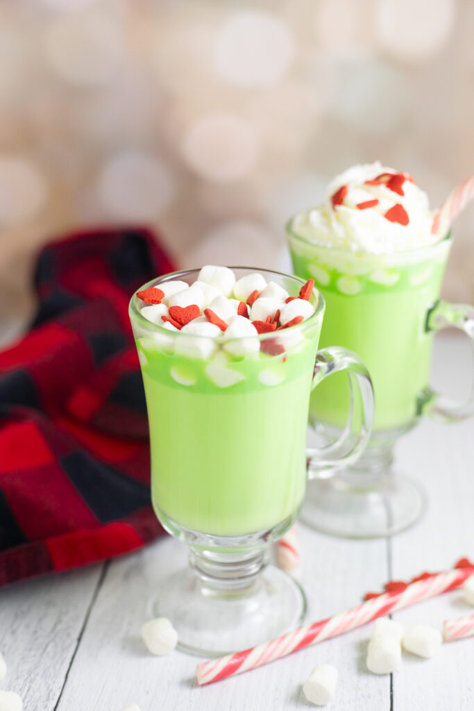 Two glass mugs with stems filled with green Grinch hot chocolate, surrounded by marshmallows, a candy cane, and red and black buffalo plaid linen napkin on a white rustic wood background. The front mug is topped with mini marshmallows and red heart sprinkles and the other mug is topped with whipped cream.