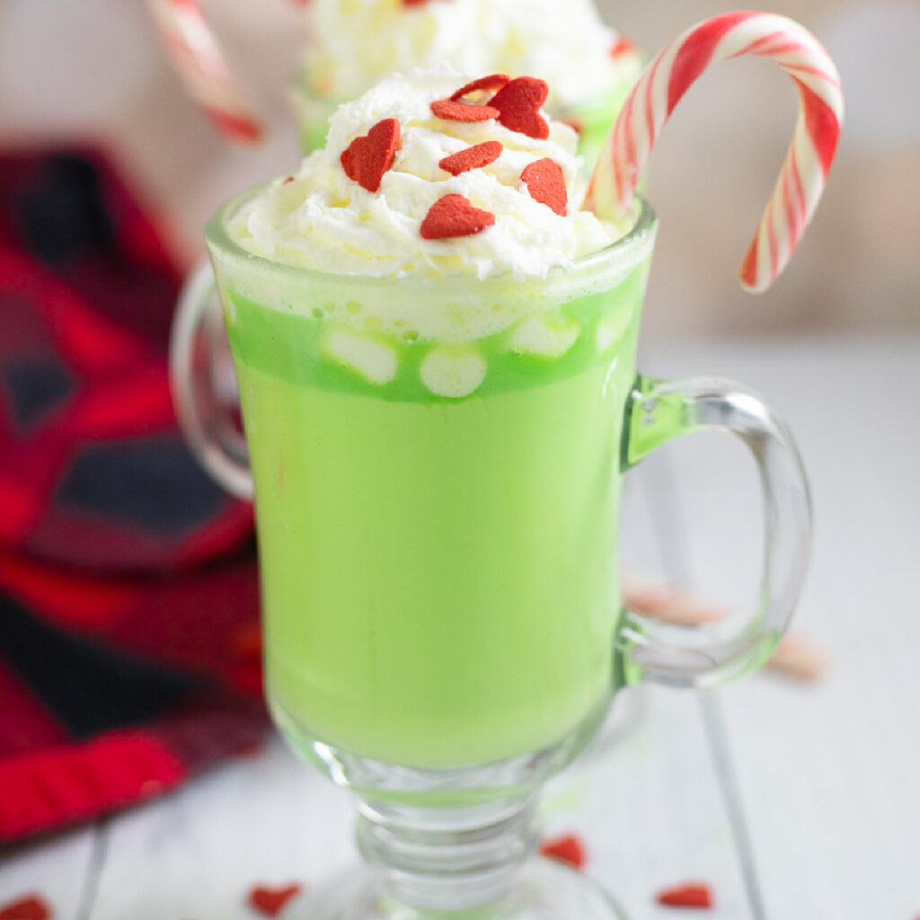 Green Grinch hot chocolate in a glass mug with a stem. The hot chocolate is topped with whipped cream, small red heart sprinkles, and a peppermint candy cane.