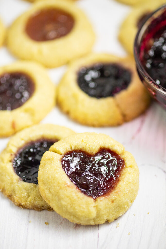 Two jam thumbprint cookies on a white background. The top cookie has a heart indent filled with red jam.