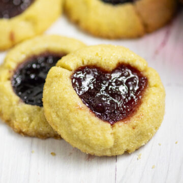 Two jam thumbprint cookies on a white background. The top cookie has a heart indent filled with red jam.