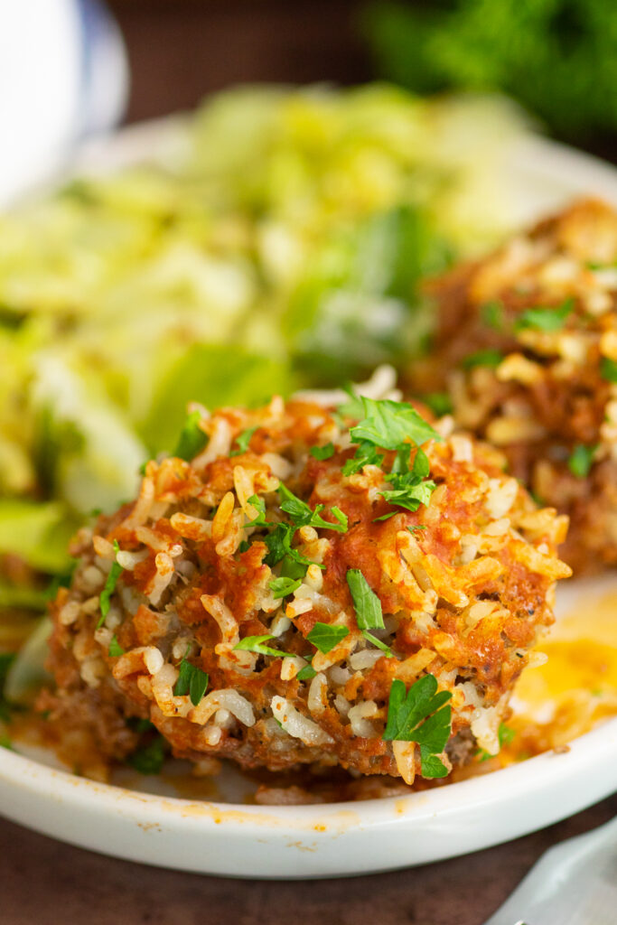 Porcupine meatball topped with red sauce and fresh parsley on a white plate with another meatball and fried cabbage behind it.