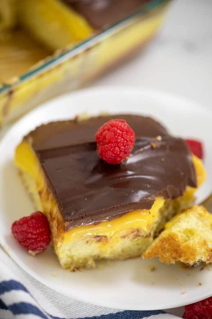 A square slice of Boston Cream poke cake on a small white plate with a bite taken out and a raspberry on top.
