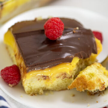 A square slice of Boston Cream poke cake on a small white plate with a bite taken out and a raspberry on top.