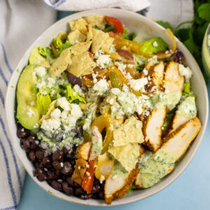 Overhead view of a chicken fajita salad in a shallow white bowl with sliced chicken, avocado, peppers and onions, black beans, crushed tortilla chips, cotija cheese, and creamy cilantro lime dressing all visible on top of the salad.