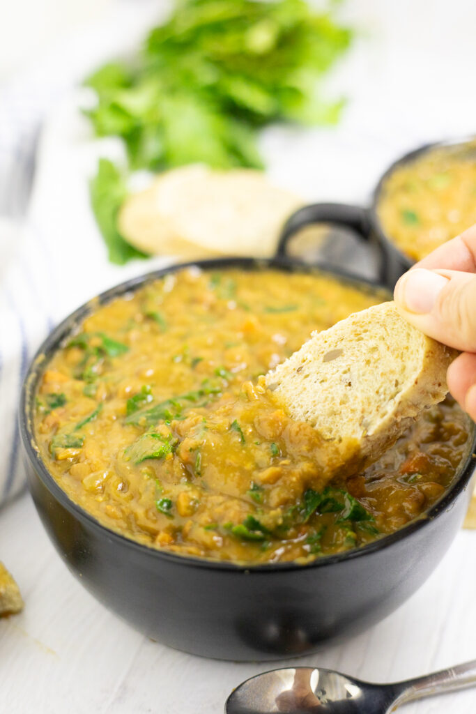 A slice of bread being dipped into a black matte bowl of instant pot lentil soup on a white wood background.