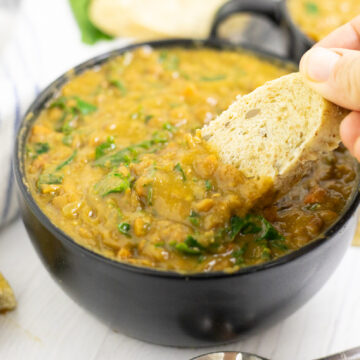 A slice of bread being dipped into a black matte bowl of instant pot lentil soup on a white wood background.