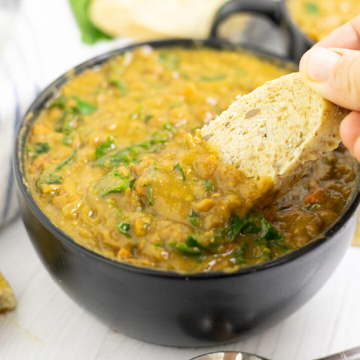 A slice of bread being dipped into a black matte bowl of instant pot lentil soup on a white wood background.