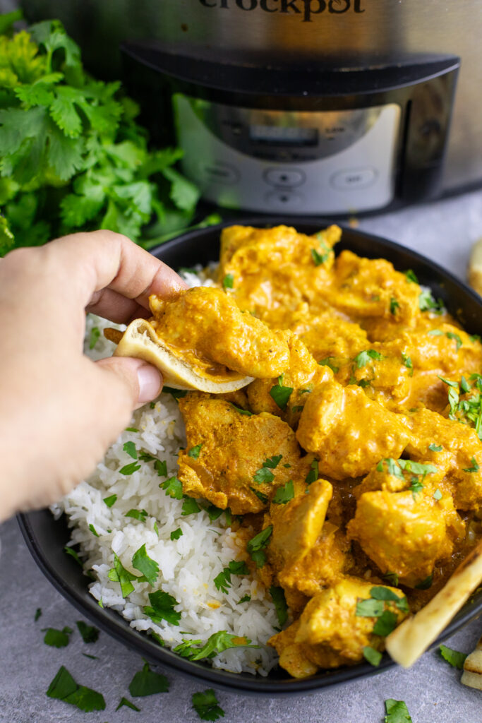 A hand picking up a piece of chicken tikka masala from a black matte bowl inside a small piece of naan.