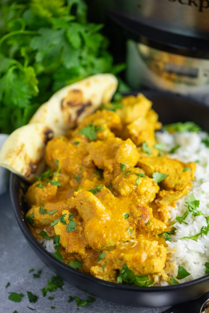 Slow Cooker Chicken Tikka Masala in a black matte bowl with white rice, topped with fresh cilantro and a piece of naan, with a bunch of fresh cilantro and the slow cooker behind.