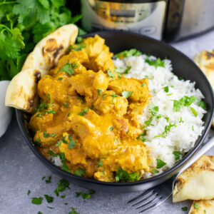 A black matte bowl with half white rice and half slow cooker chicken tikka masala, topped with fresh cilantro and a piece of naan.