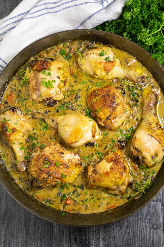 Overhead view of chicken fricassee in a large cast iron skillet topped with fresh thyme and surrounded by fresh parsley and a linen napkin.