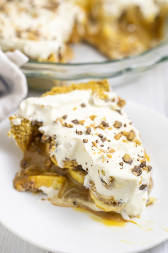 A slice of banoffee pie topped with toffee bits on a white plate with the remaining pie in a glass pie dish behind.