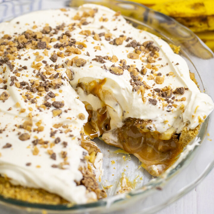 Banoffee pie in a glass pie dish with a slice cut out showing the gooey interior. The pie is topped with homemade whipped cream and chocolate toffee bits with a bunch of bananas behind.