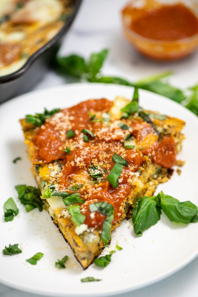 A slice of pizza frittata on a small white plate topped with pizza sauce and freshly grated parmesan next to basil leaves with a cast iron skillet and a small glass bowl of pizza sauce behind.