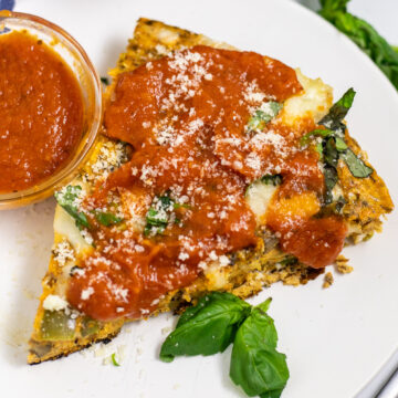 A slice of pizza frittata on a small white plate topped with pizza sauce and freshly grated parmesan next to a small glass bowl of pizza sauce and basil leaves.