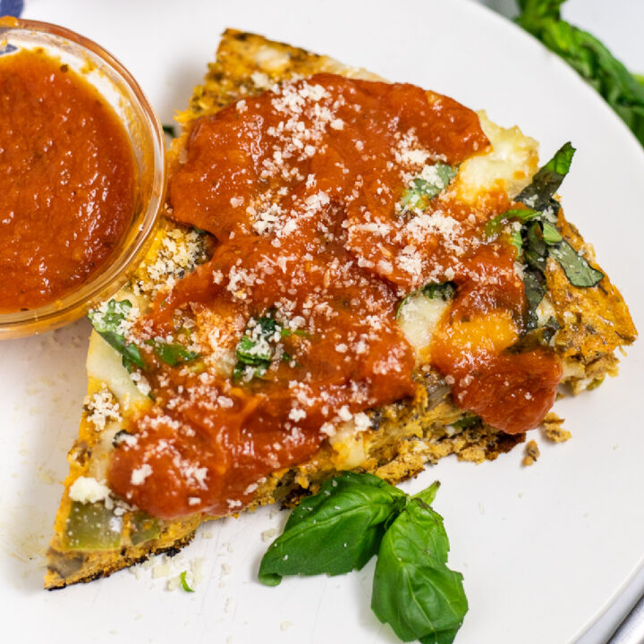 A slice of pizza frittata on a small white plate topped with pizza sauce and freshly grated parmesan next to a small glass bowl of pizza sauce and basil leaves.