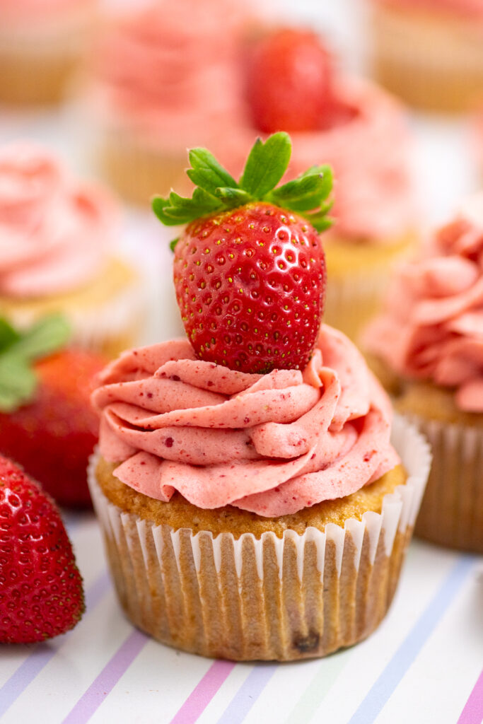 Strawberry cupcake topped with strawberry frosting and a fresh strawberry, surrounded by more fresh strawberries and cupcakes on a pastel striped background.
