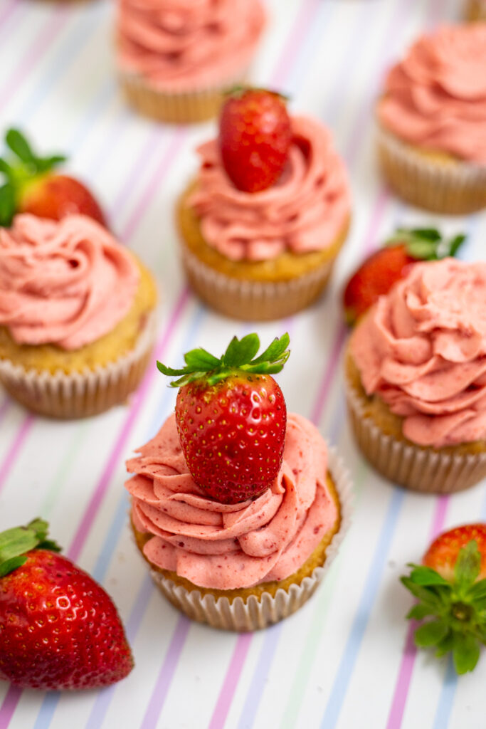Overhead view of strawberry cupcakes topped with strawberry frosting and fresh strawberries on a pastel striped background with fresh strawberries scattered around.