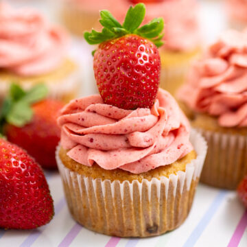 Strawberry cupcake topped with strawberry frosting and a fresh strawberry, surrounded by more fresh strawberries and cupcakes on a pastel striped background.